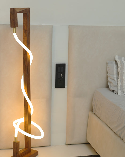 Sculpted glow wooden floor lamp with a warm LED rope curving through an open solid wood frame, standing beside a bed