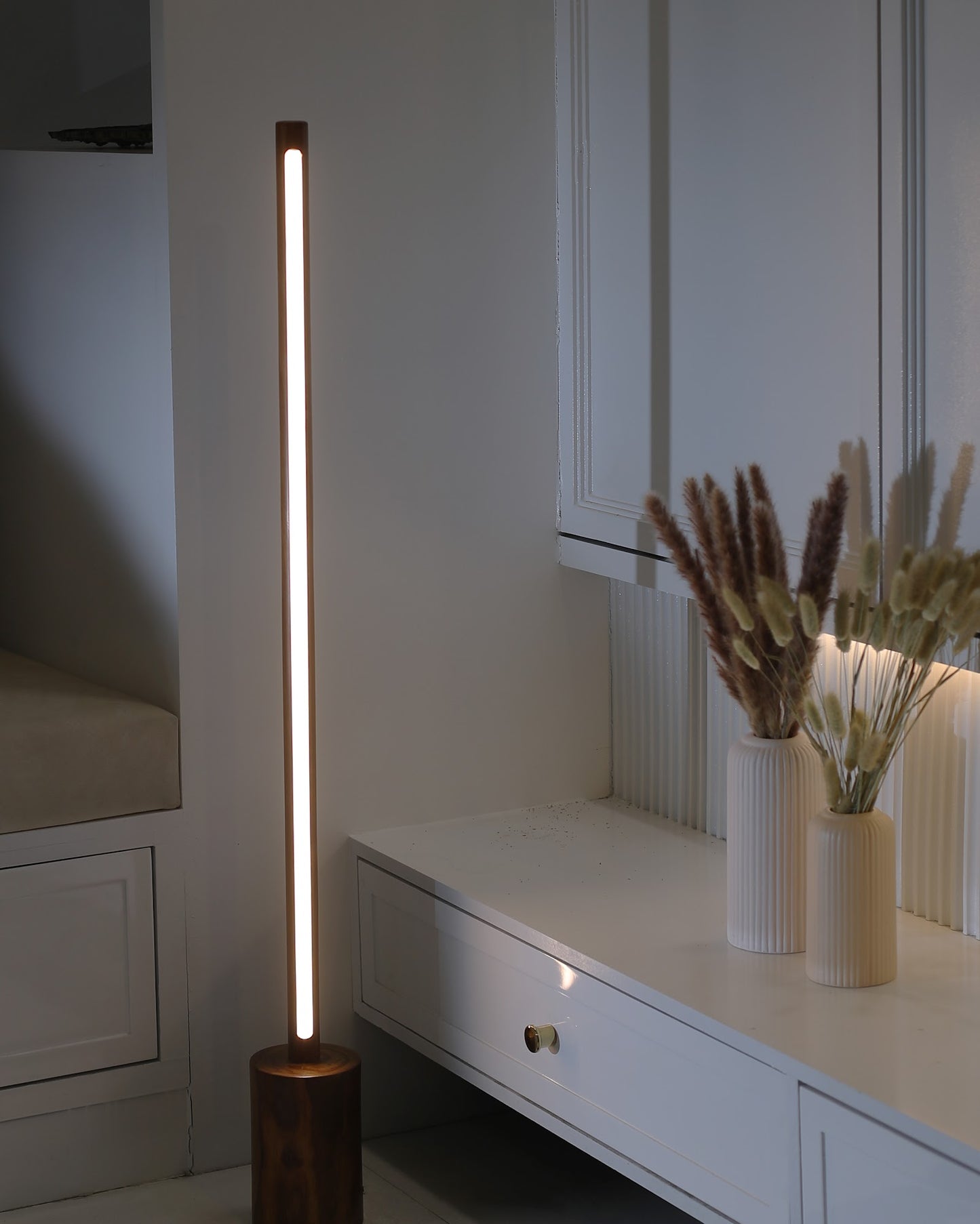 Minimal tall wooden floor lamp with slim cylindrical rod lit warm white, standing on a round turned solid wood base beside a white console table