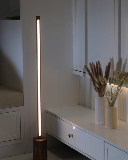 Minimal tall wooden floor lamp with slim cylindrical rod lit warm white, standing on a round turned solid wood base beside a white console table