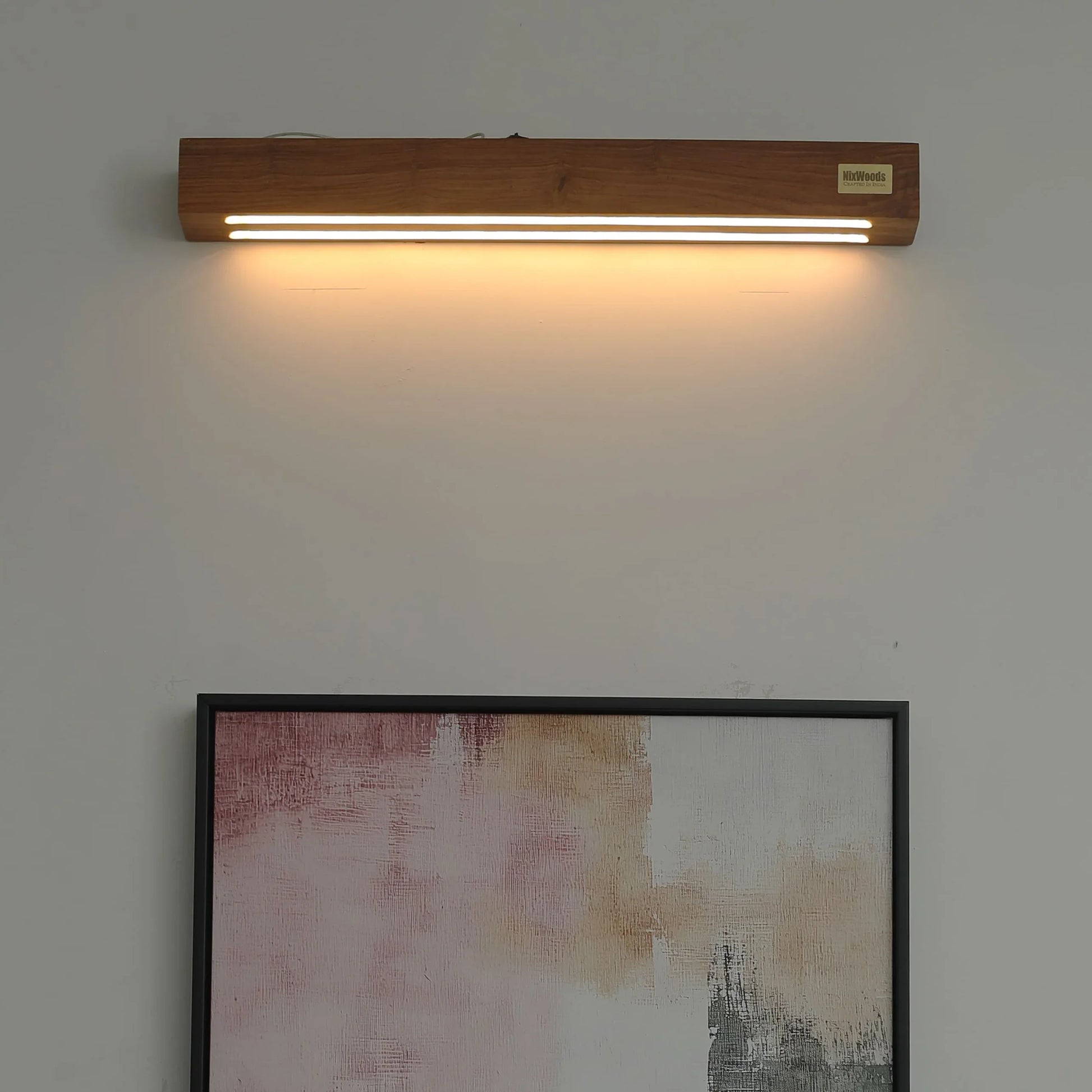 Rosewood linear wall lamp lit above framed art on a living room wall - NixWoods solid wood wall sconce