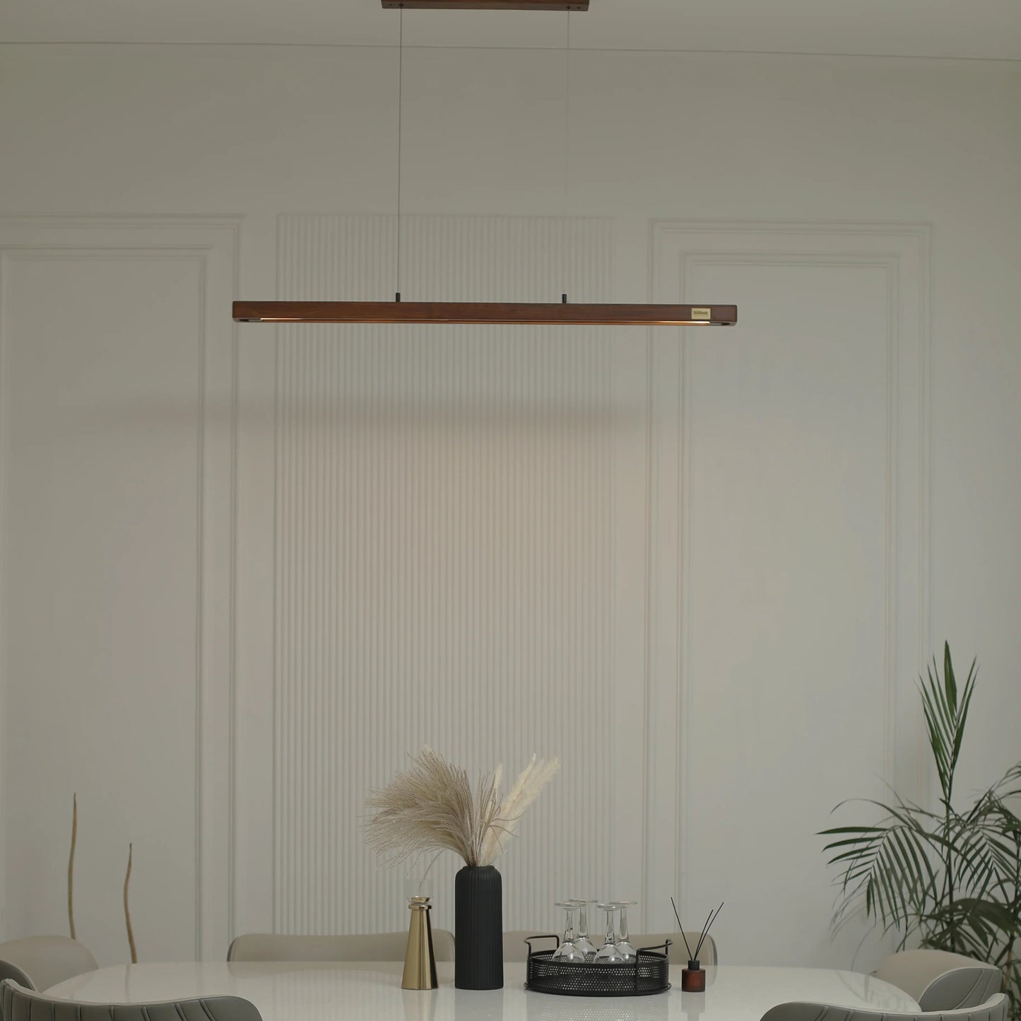 A modern dining room setup featuring a white marble-top dining table with a sculptural curved wooden base, surrounded by gray upholstered wooden chairs. A long horizontal linear pendant light hangs above the table, and a potted plant is visible on the right against a white paneled wall.
