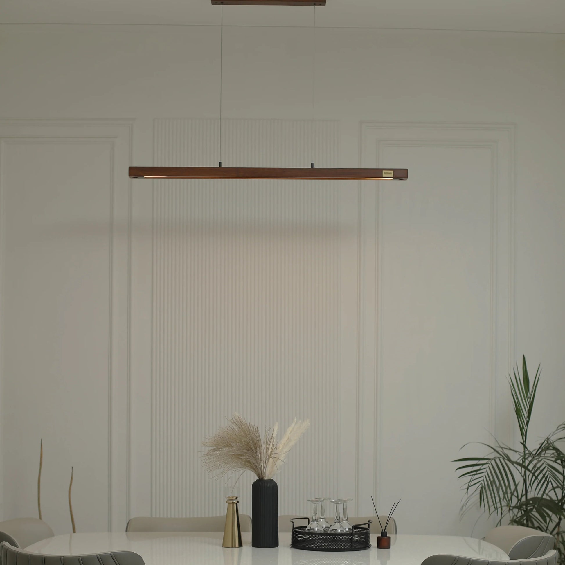 A modern dining room setup featuring a white marble-top dining table with a sculptural curved wooden base, surrounded by gray upholstered wooden chairs. A long horizontal linear pendant light hangs above the table, and a potted plant is visible on the right against a white paneled wall.