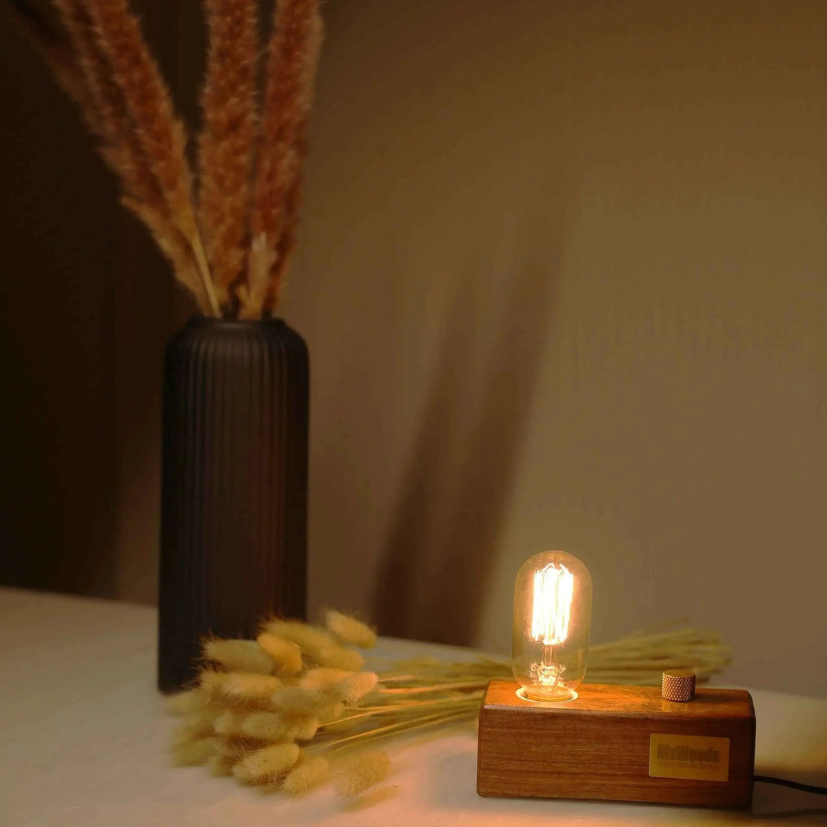 Rosewood block lamp with Edison bulb lit beside a black vase of dried pampas grass