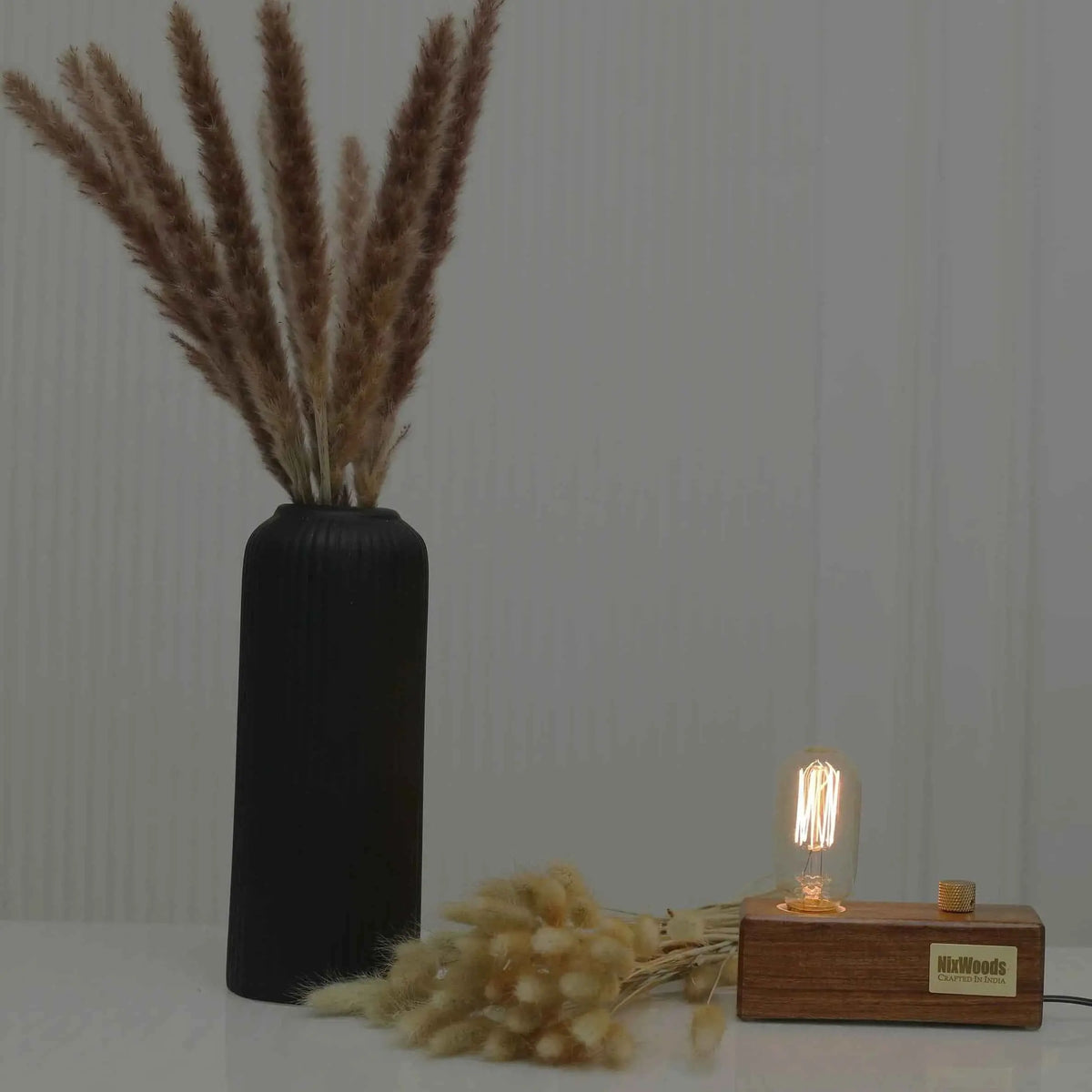 Rosewood Edison bulb lamp on a white table beside a black ribbed vase and bunny tails