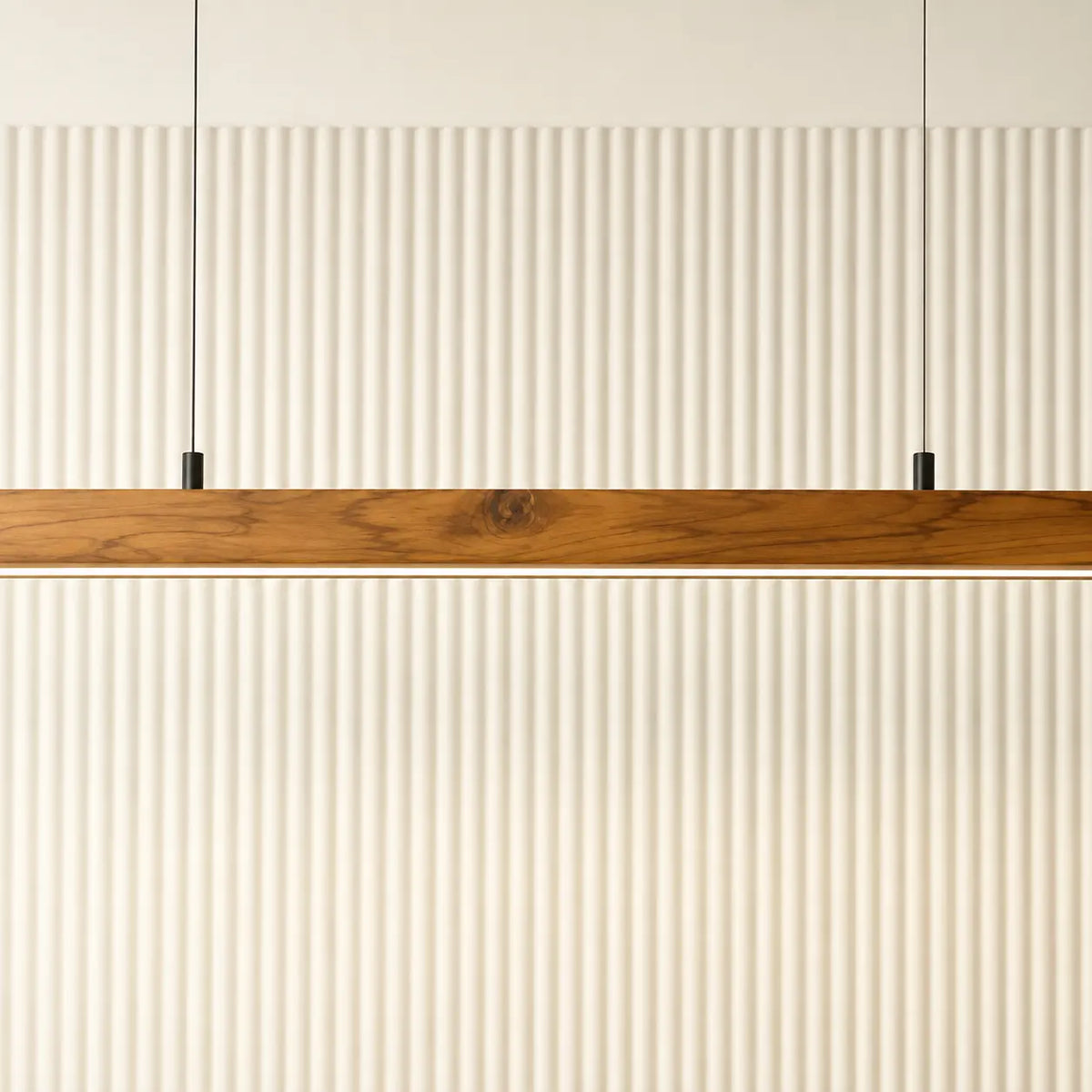 Teak linear pendant bar suspended on slim black cables in front of a fluted cream wall