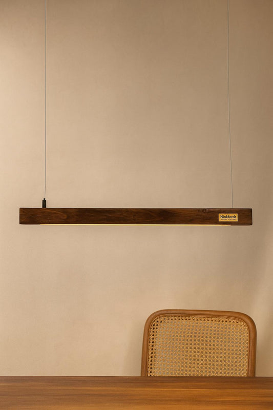 Long rectangular wooden light fixture on a beige wall above a wooden table with a wicker chair.